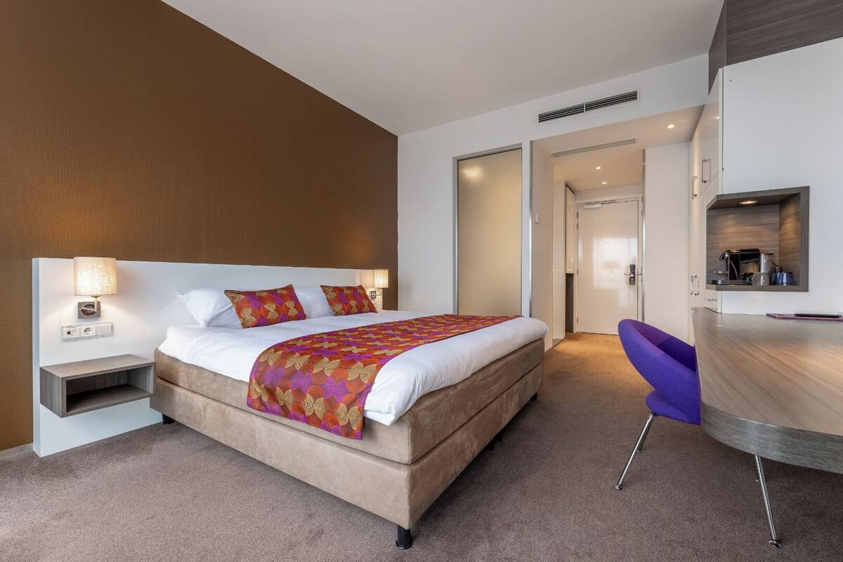 A modern hotel room with a large bed, a desk with a chair, and a small kitchenette.