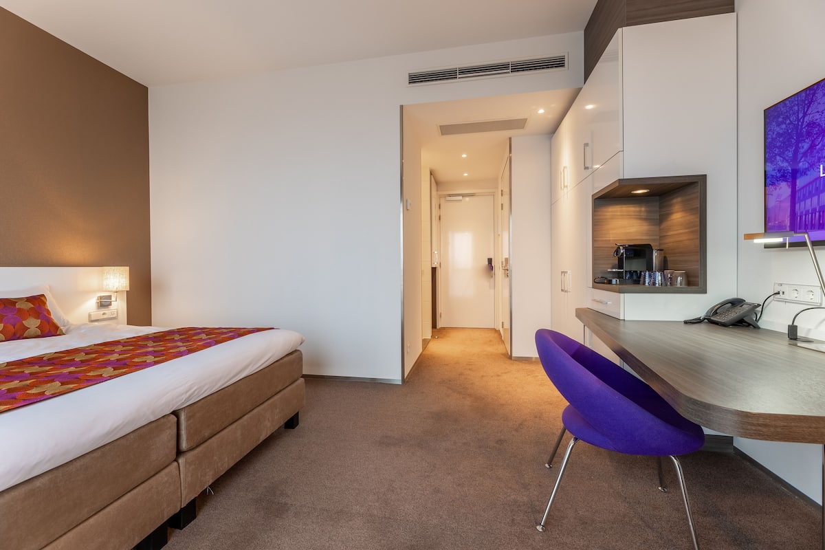 A modern hotel room with a bed, a desk, a chair, and a kitchenette.