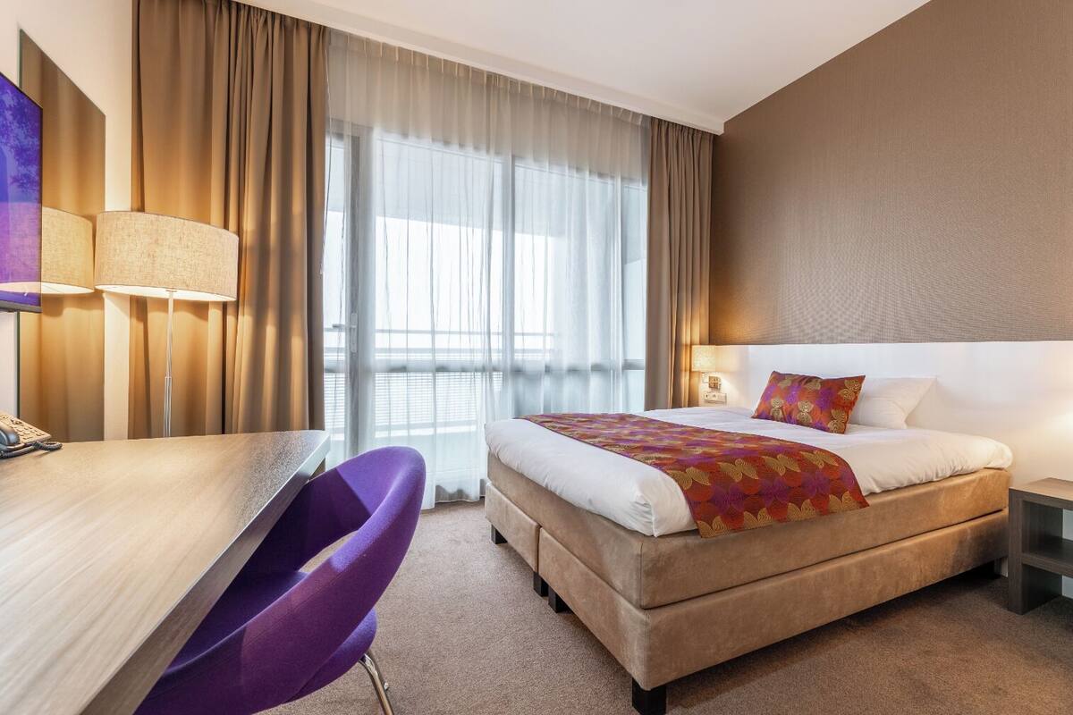 A hotel room with a bed, desk, chair, and a large window with curtains.