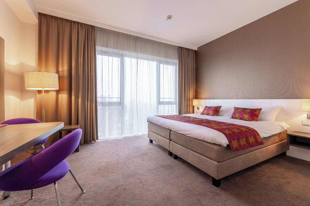 A hotel room with a bed, a desk, a chair, and a large window with curtains.