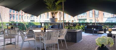An outdoor patio with white metal chairs and tables, a large potted plant, and a black umbrella providing shade.