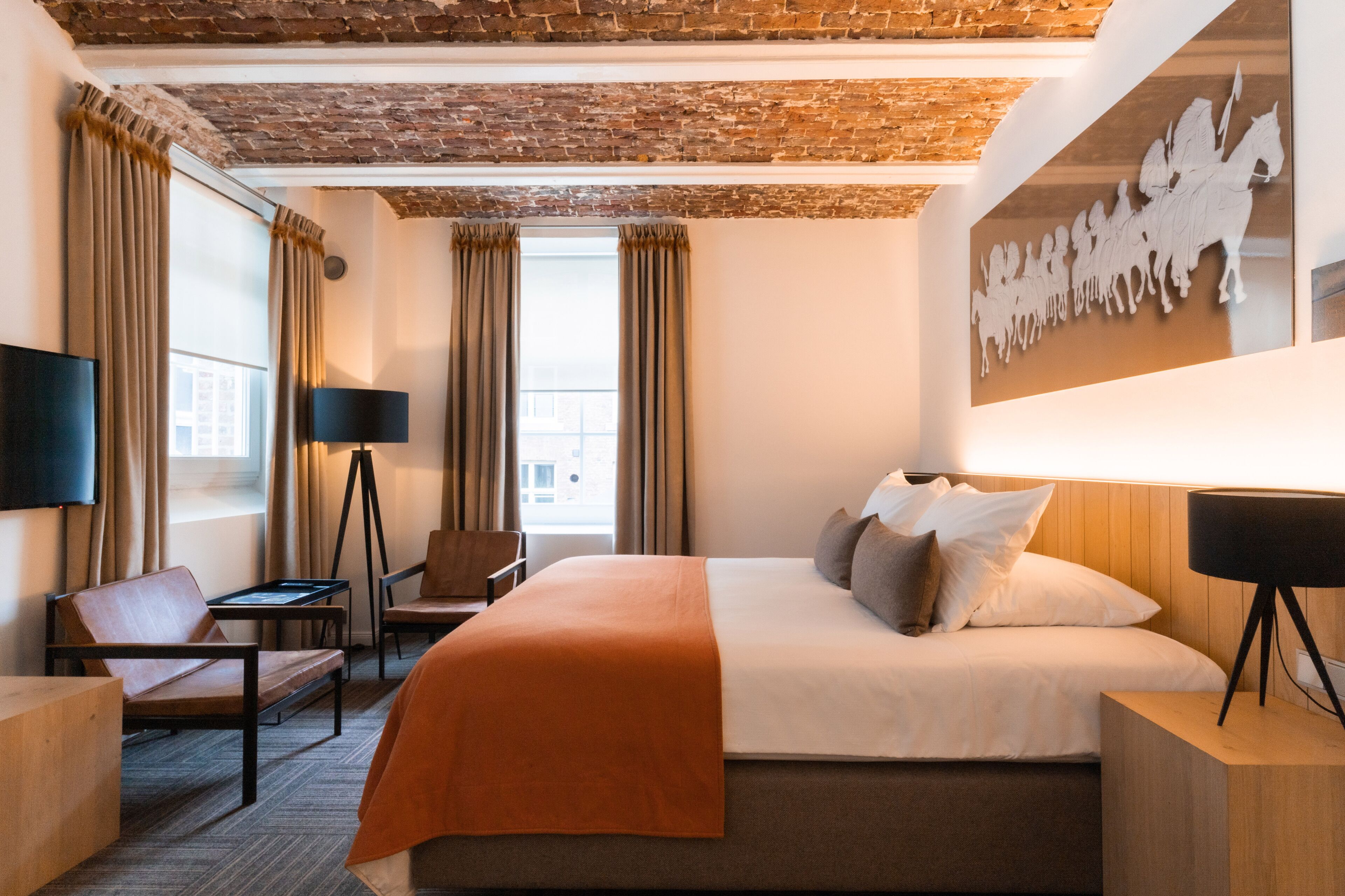 A hotel room with a brick ceiling, a large bed with an orange throw, a flat-screen TV, a desk, and a chair.