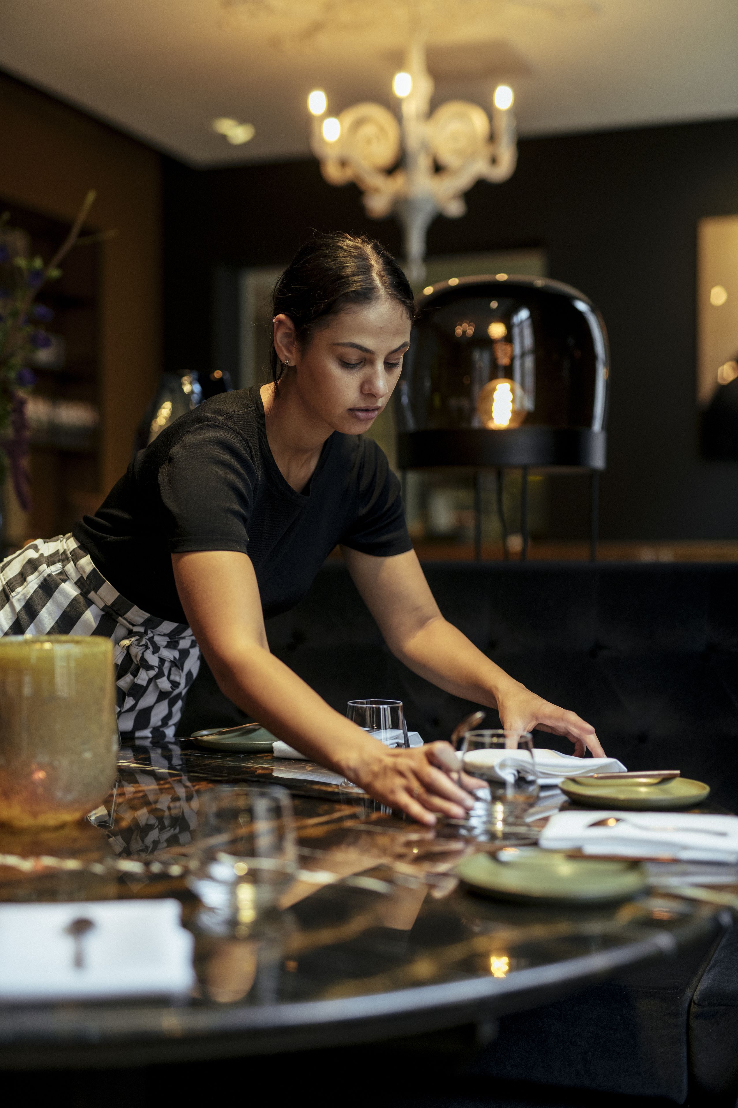 Une personne qui dresse une table avec des assiettes et des verres, un lustre et un décor intérieur moderne.