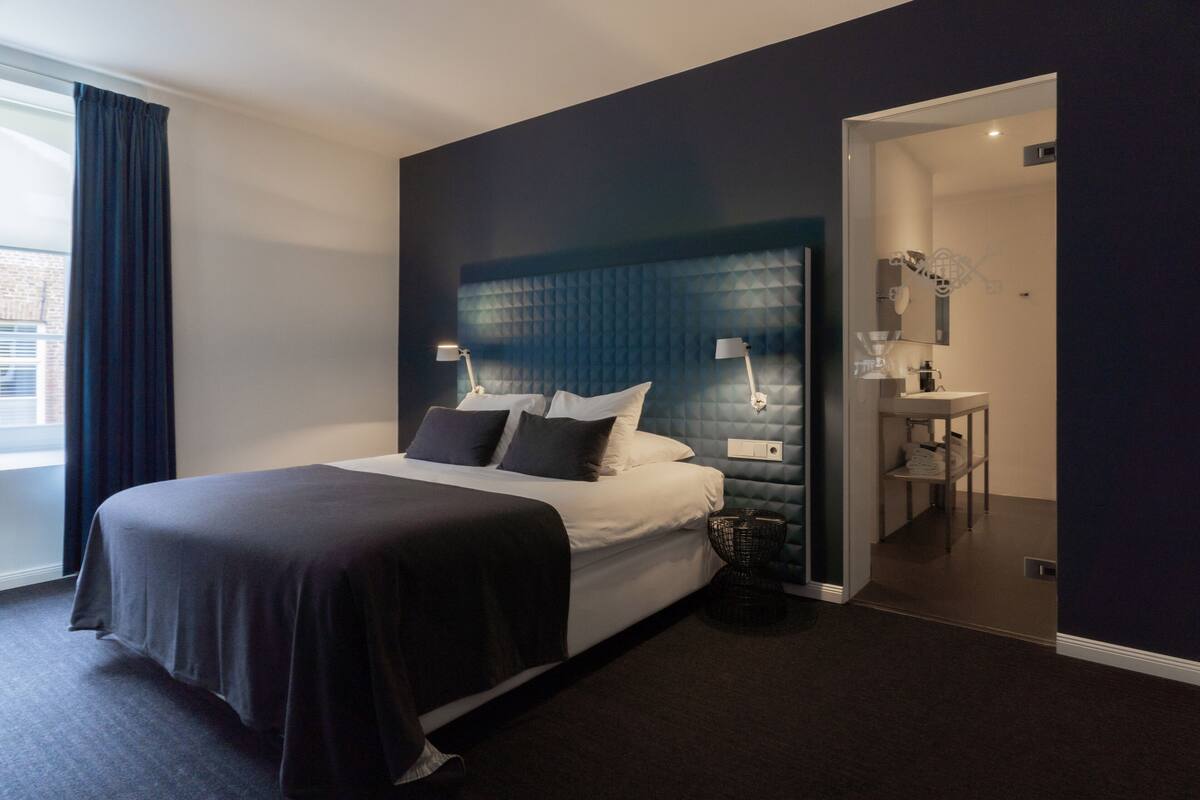 Ein modernes Hotelzimmer mit einem großen Bett, Nachttischlampen, einem dunklen Kopfteil, einem kleinen Beistelltisch und Blick auf die Stadt durch ein Fenster.