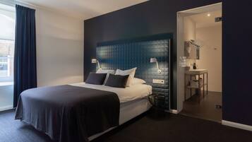 A modern hotel room with a large bed, bedside lamps, a dark headboard, a small side table, and a view of the city through a window.