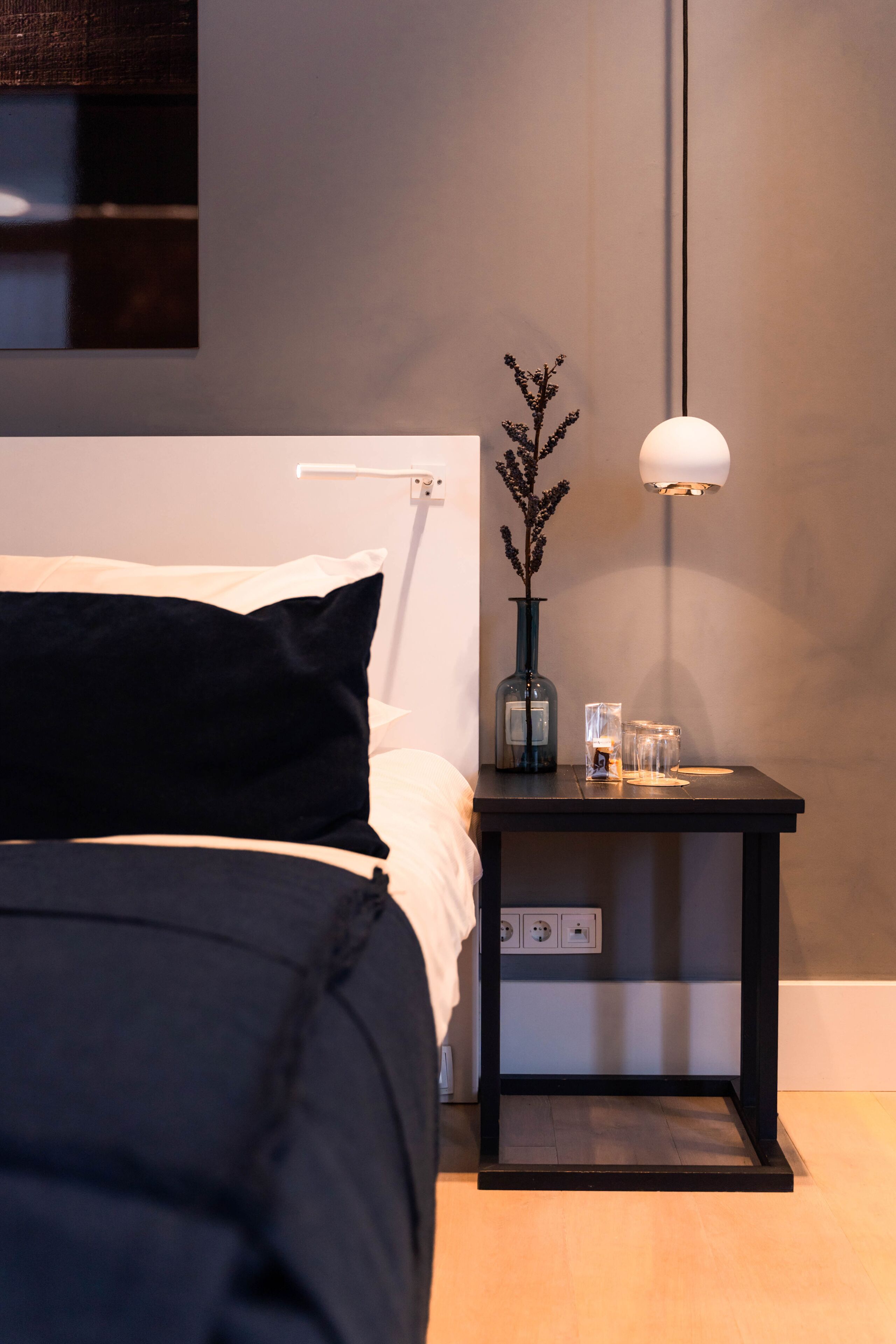 Ein ordentlich bezogenes Bett mit schwarzem Kopfteil, ein Nachttisch mit einer Vase und einer Lampe sowie eine Wandleuchte.