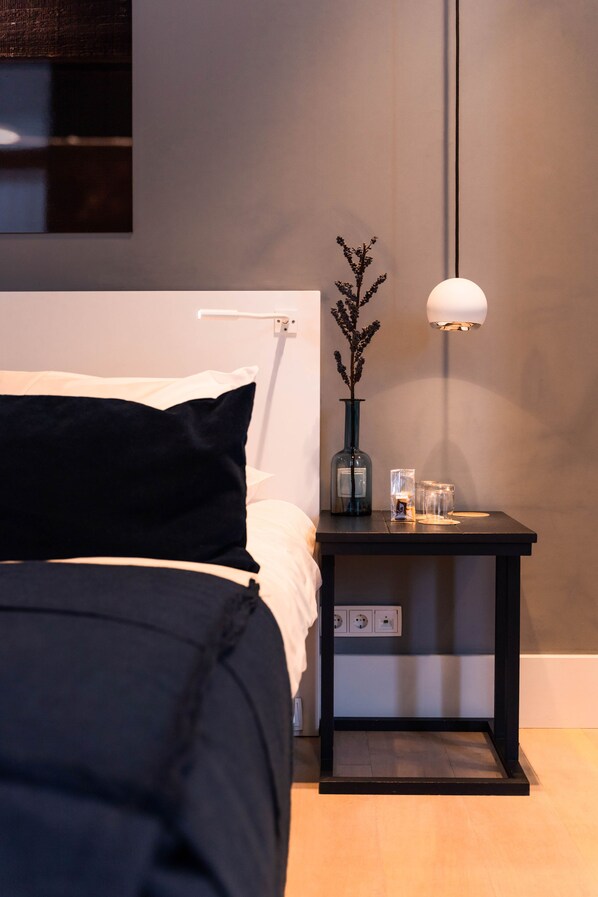 Una cama bien hecha con un cabecero negro, una mesita de noche con un florero y una lámpara, y un aplique en la pared.