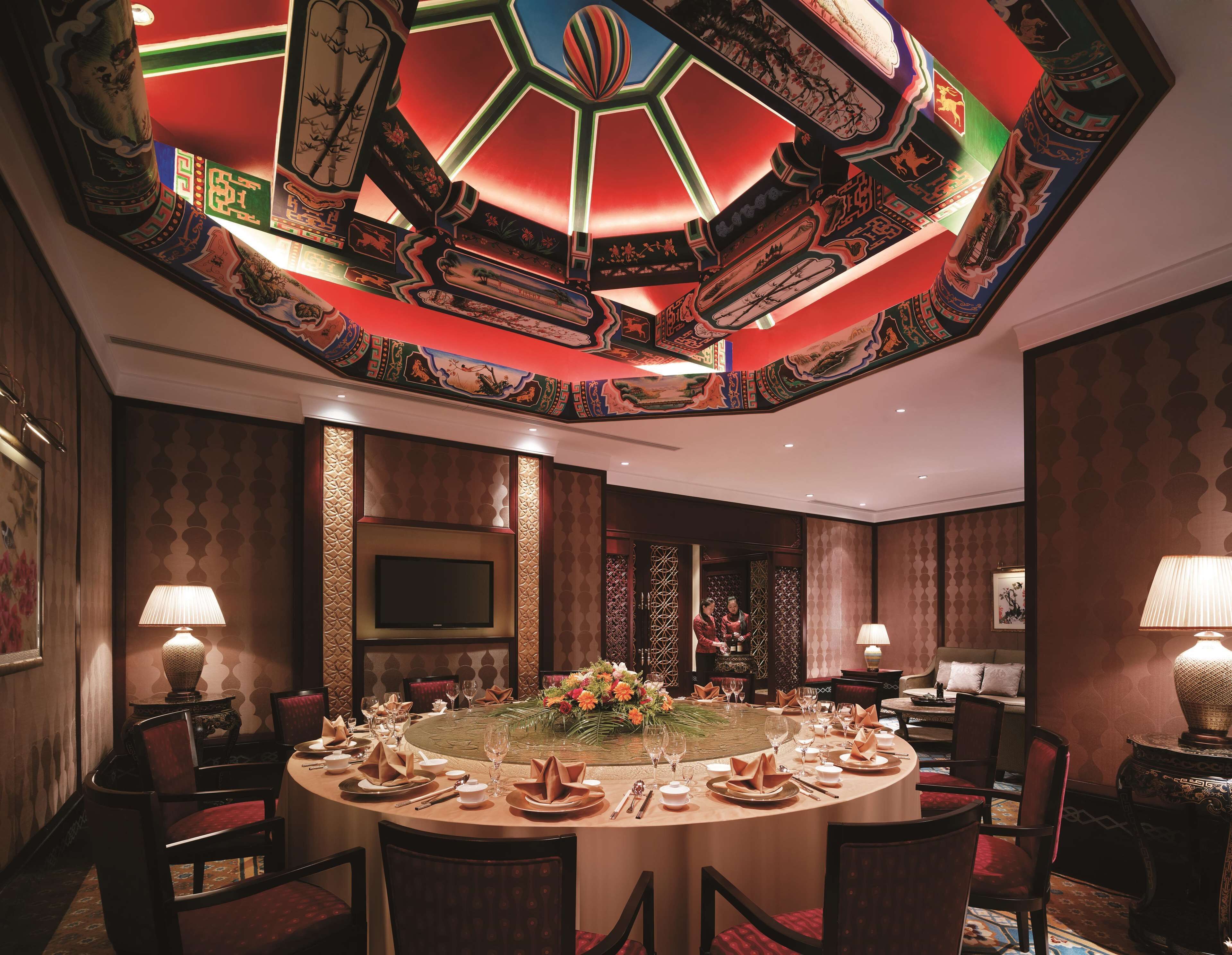Une table de salle à manger ronde, dressée pour un repas, entourée de chaises, avec un plafond décoratif et des motifs traditionnels sur les murs.