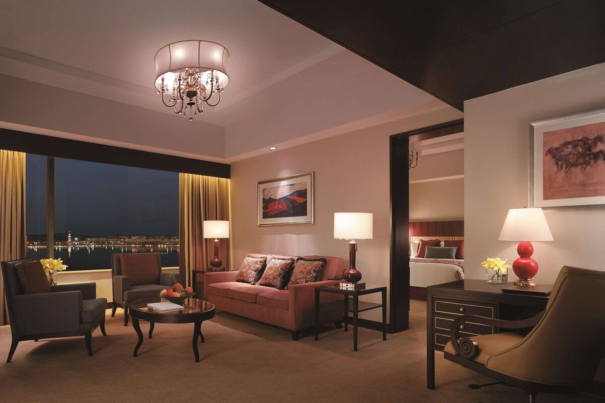 Une chambre d’hôtel avec un canapé, des fauteuils, une table basse, un bureau et une lampe. Un tableau orne le mur et la fenêtre offre une vue sur la ville la nuit.