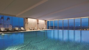 Ein modernes Hallenbad mit klarem, blauem Wasser, großen Fenstern mit Blick auf die Stadt und Liegestühlen am Beckenrand.