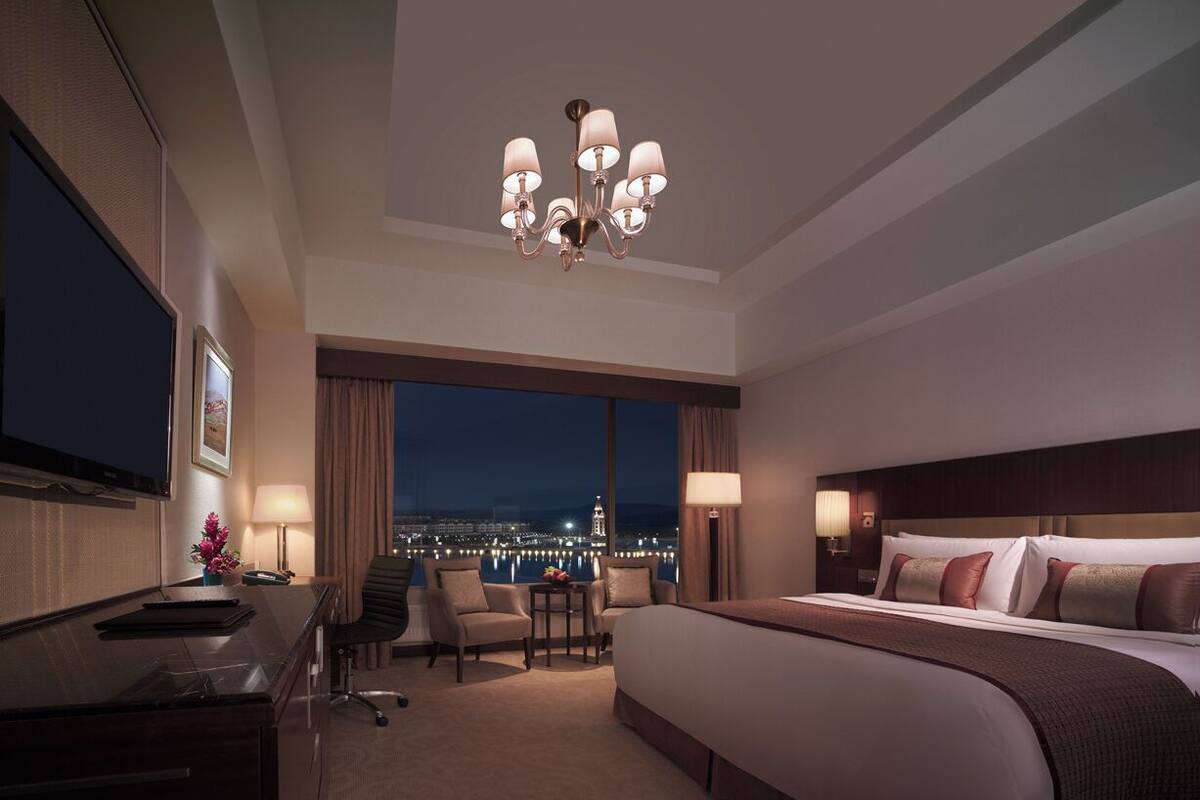 Une chambre d’hôtel avec un grand lit, un bureau, une chaise, une petite table et une vue sur la ville la nuit.