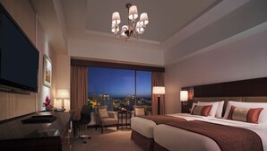 Ein Hotelzimmer mit einem großen Bett, einem Schreibtisch, einem Sessel, einem Fernseher und Blick auf die nächtliche Stadt.