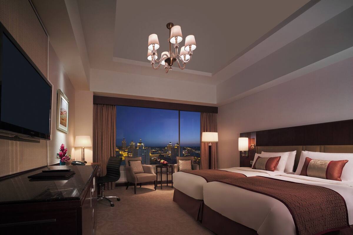 Une chambre d’hôtel avec un grand lit, un bureau, une chaise, une télévision et une vue sur la ville la nuit.