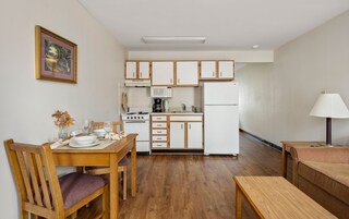 Una cocina compacta con armarios blancos, estufa, microondas y refrigerador. Hay una mesa de comedor con sillas y una mesita con una lámpara.