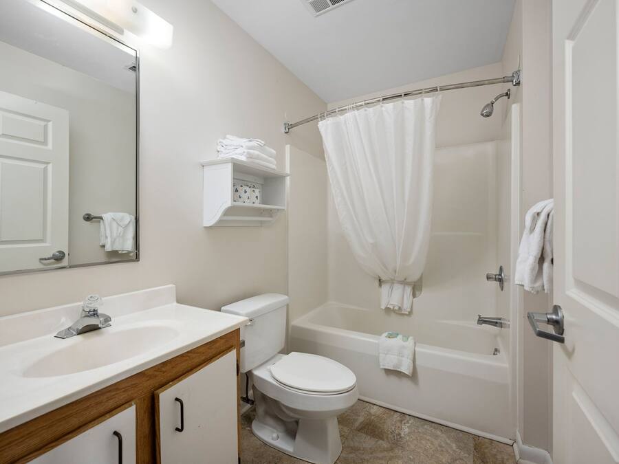Un bany amb un lavabo, vàter i banyera de color blanc. Hi ha un mirall sobre el lavabo i un penjador de tovalloles sobre la banyera.