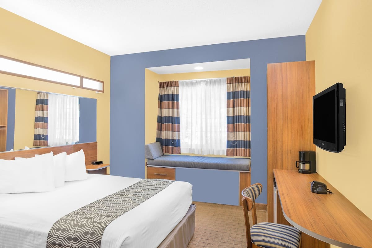 A hotel room with a bed, a desk, a television, a window with curtains, and a small seating area.