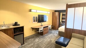 A compact hotel room with a kitchenette, a flat-screen TV, a sofa, and a small bed.