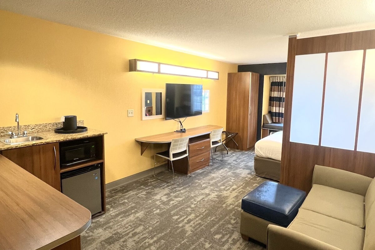 A compact hotel room with a kitchenette, a flat-screen TV, a sofa, and a small bed.