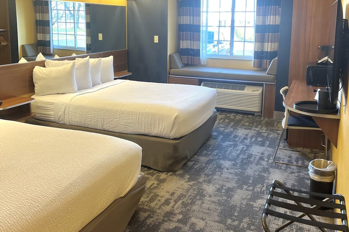 A hotel room with two beds, a desk, a chair, a TV, and a window with curtains.