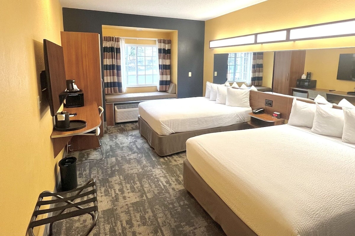 A hotel room with two beds, a desk with a chair, a small table, a trash bin, and a window with curtains.