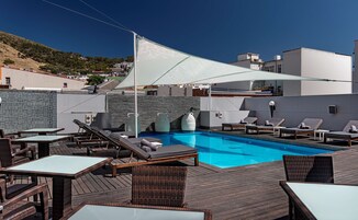 A rooftop pool area with lounge chairs and tables, a clear blue pool, and a view of the surrounding buildings and hills.