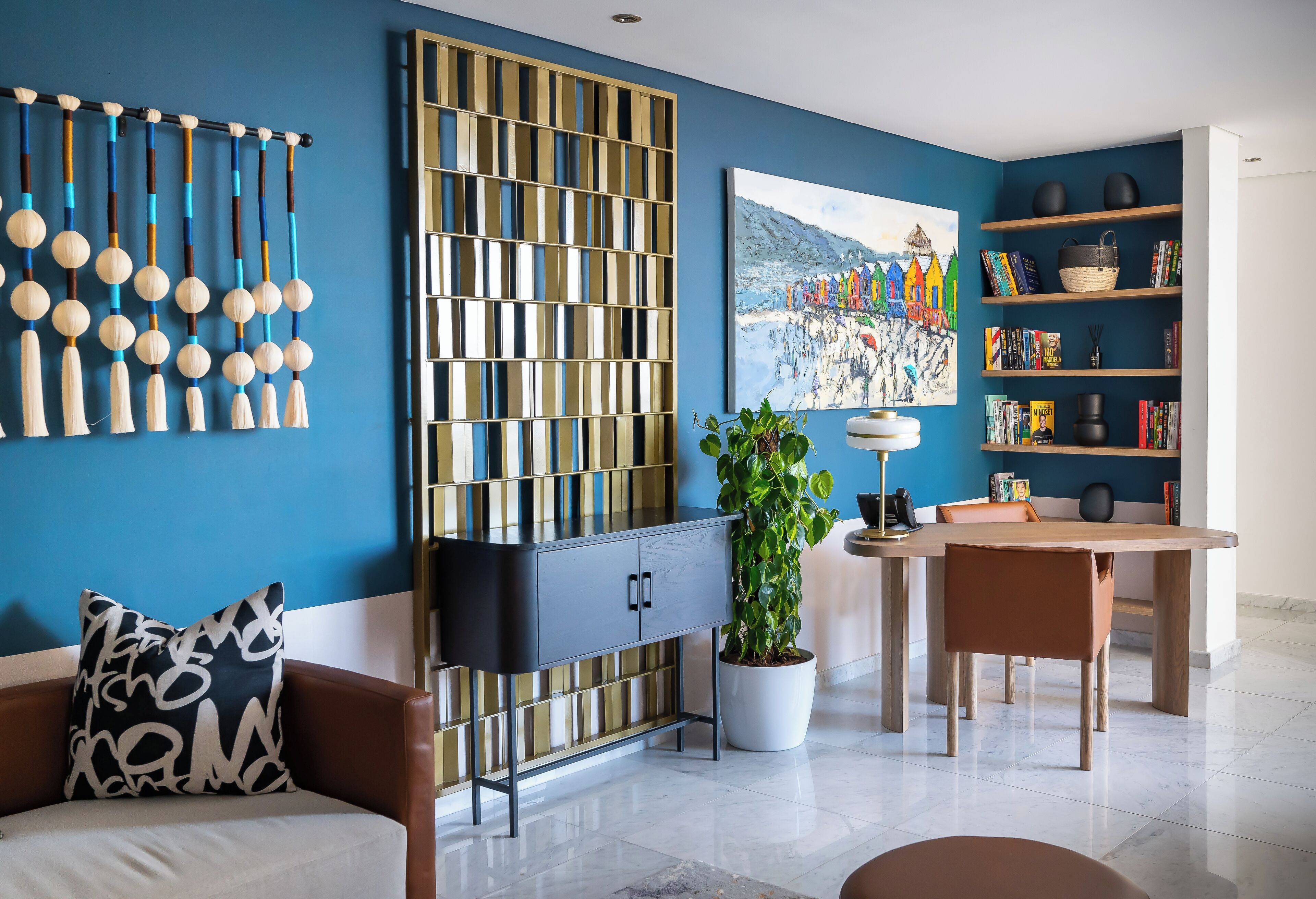 A modern living room with a blue wall, a decorative wall hanging, a bookshelf, a dining area, and a potted plant.