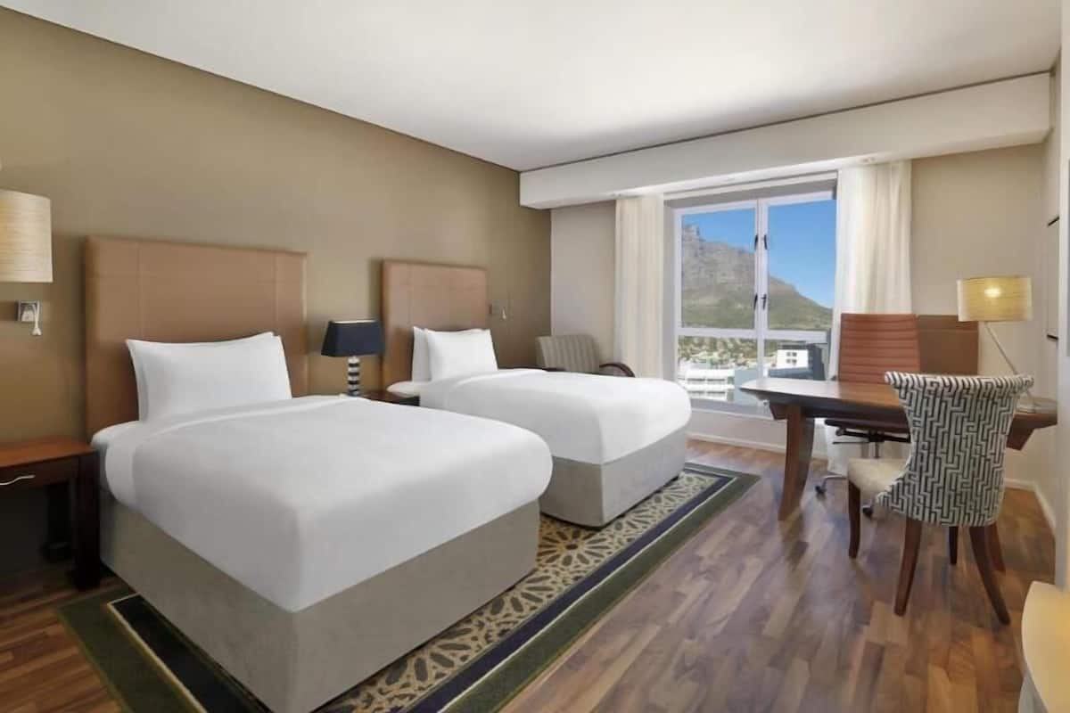 Une chambre d’hôtel avec deux lits, un bureau, une chaise, une fenêtre donnant sur un paysage agréable et un tapis à motifs.