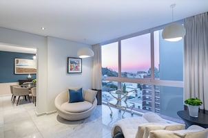 A modern living room with a large window, a round sofa, a dining area, and a view of the cityscape.