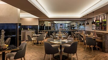 A modern restaurant with a central bar, multiple dining tables, and a herringbone wood floor.
