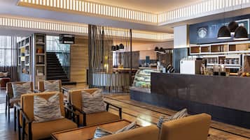 Un moderno vestíbulo de hotel con zonas de estar, una escalera, una cafetería y vistas al exterior.