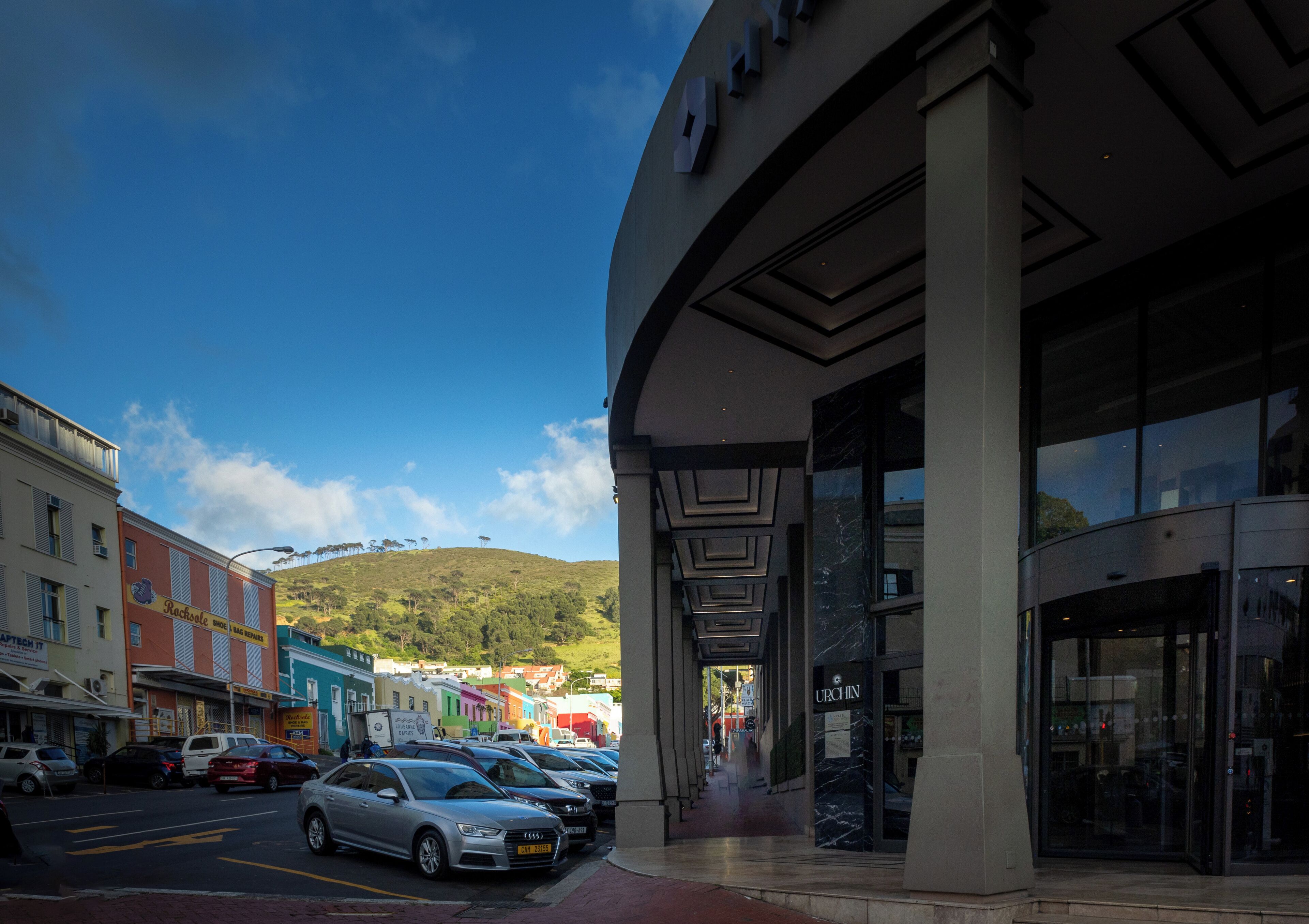 A street view with parked cars, a building with a modern facade, and a clear blue sky.