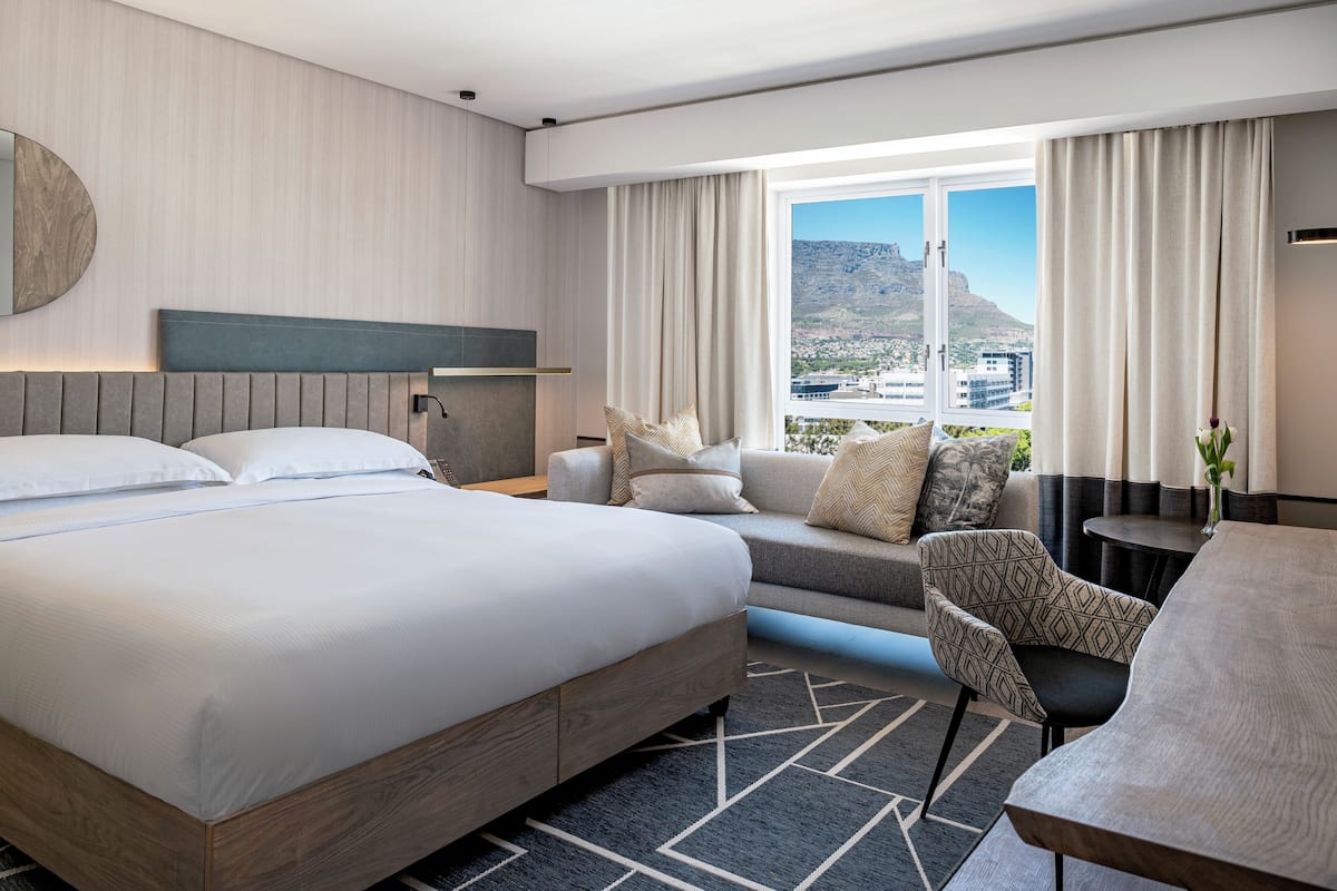 Une chambre d’hôtel moderne dotée d’un grand lit, d’un canapé, d’une table à manger et offrant une vue sur le paysage urbain grâce à de grandes fenêtres.