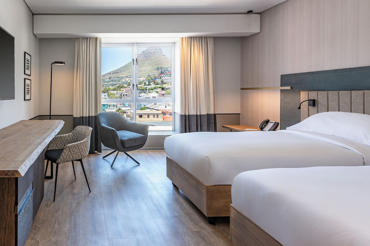 Une chambre d’hôtel avec deux lits, un bureau, une chaise et une grande fenêtre donnant sur la ville et une montagne.