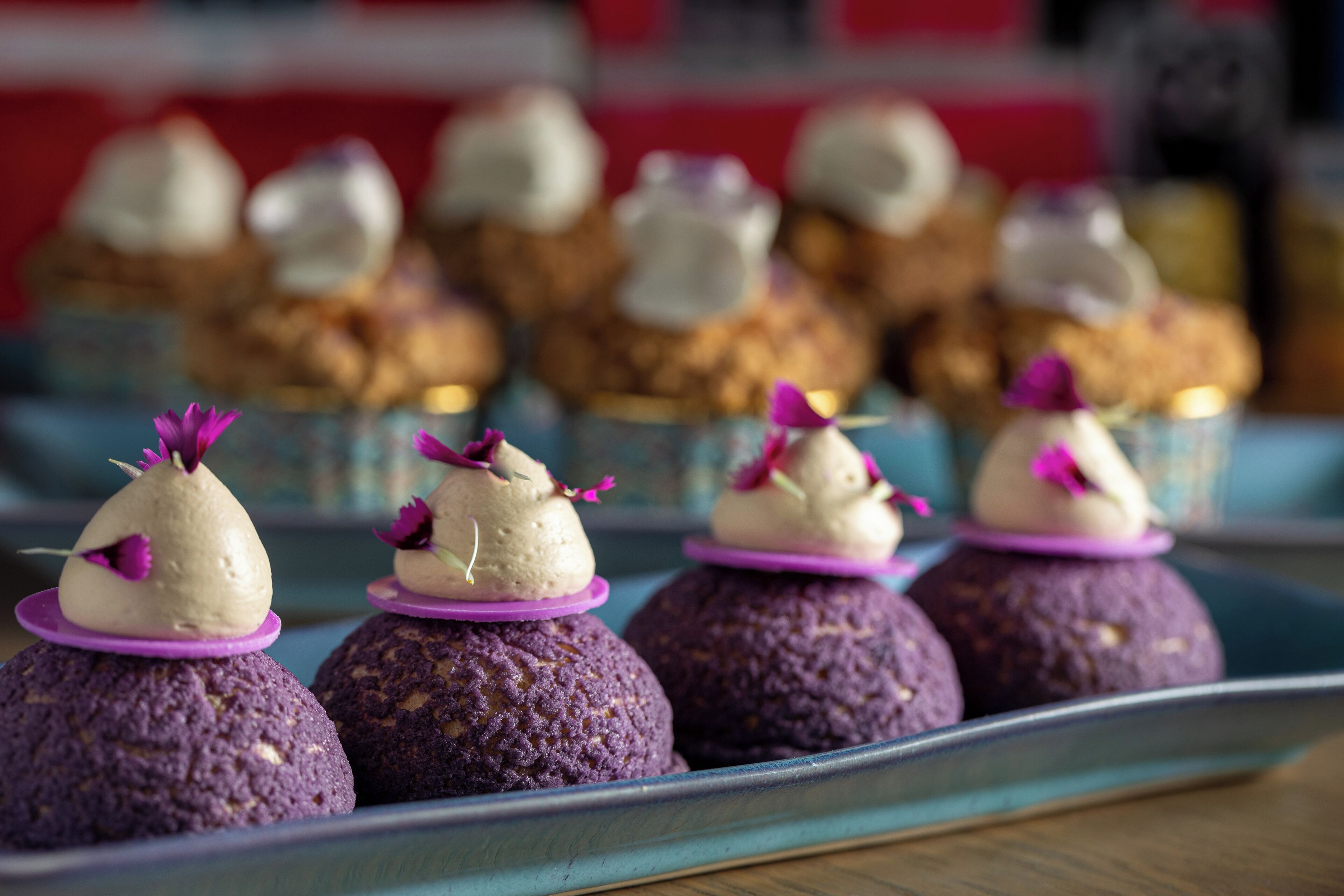 Eine Schale mit violetten Desserts, bedeckt mit weißer Sahne und verziert mit violetten Blüten, mit unscharfem Hintergrund ähnlicher Desserts.