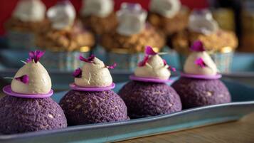 A tray of purple-colored desserts with white cream on top, garnished with purple flowers, and a blurred background of similar desserts.