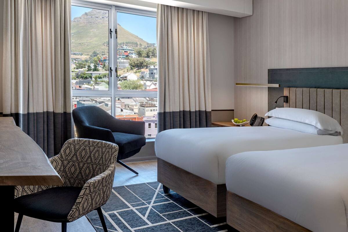 Une chambre d’hôtel avec deux lits, une table à manger, une chaise et une vue sur le paysage urbain, grâce à de grandes fenêtres.