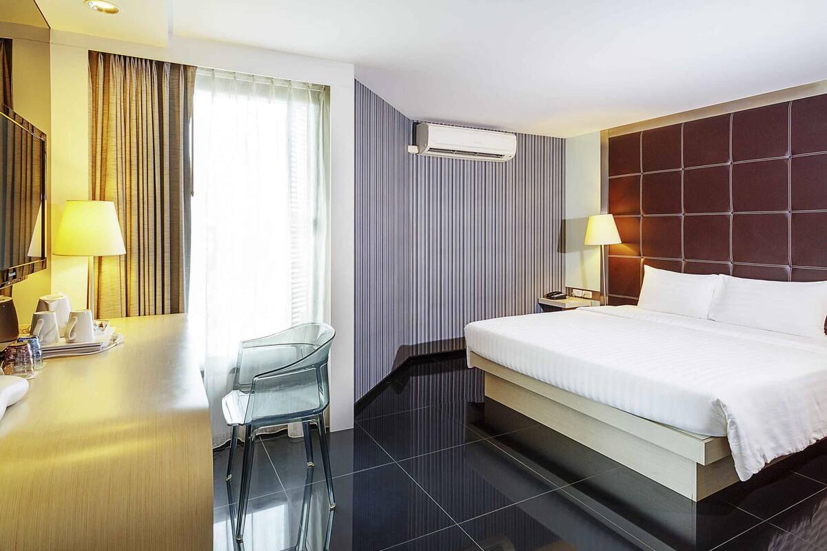Une chambre d’hôtel comprenant un lit, des tables de chevet, un bureau avec une chaise, un appareil de climatisation et une fenêtre avec des rideaux.