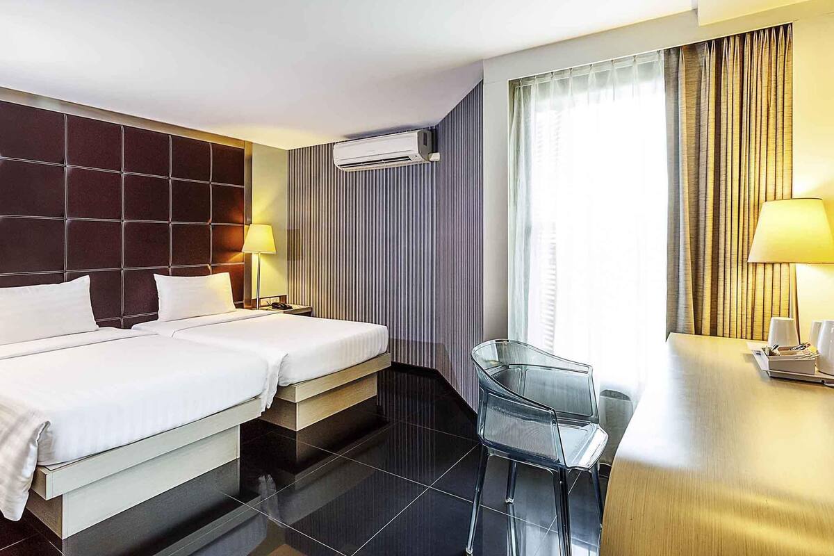 A hotel room with two beds, a desk, a chair, an air conditioning unit, and a window with curtains.