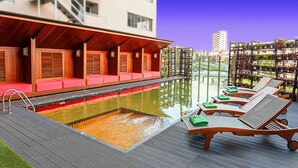 A pool area with red cushioned seating and sun loungers, a water feature, and a view of a cityscape. - ibis Styles Chiang Mai (Chiang Mai)