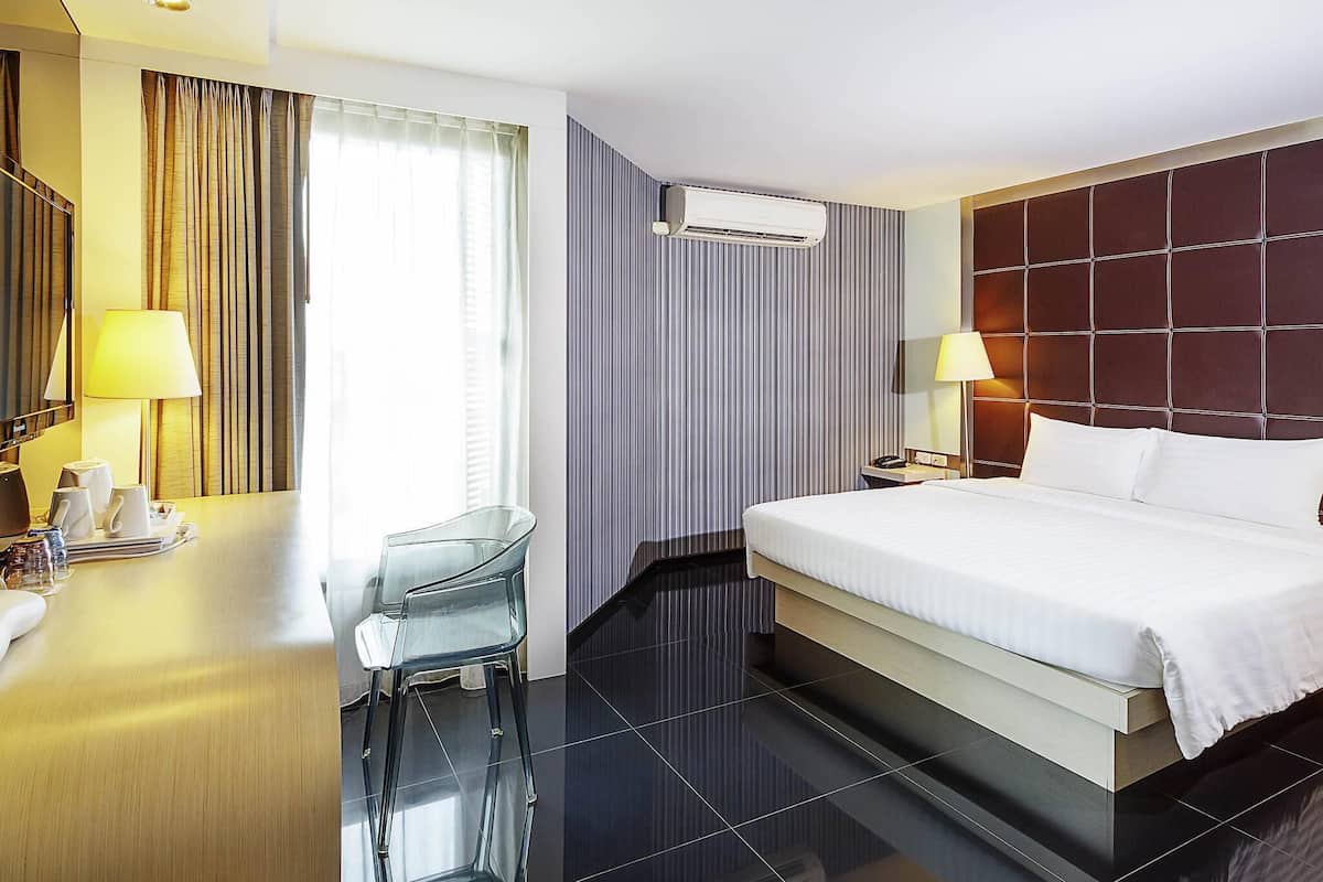 A hotel room with a bed, a desk with a chair, an air conditioning unit, and a window with curtains.