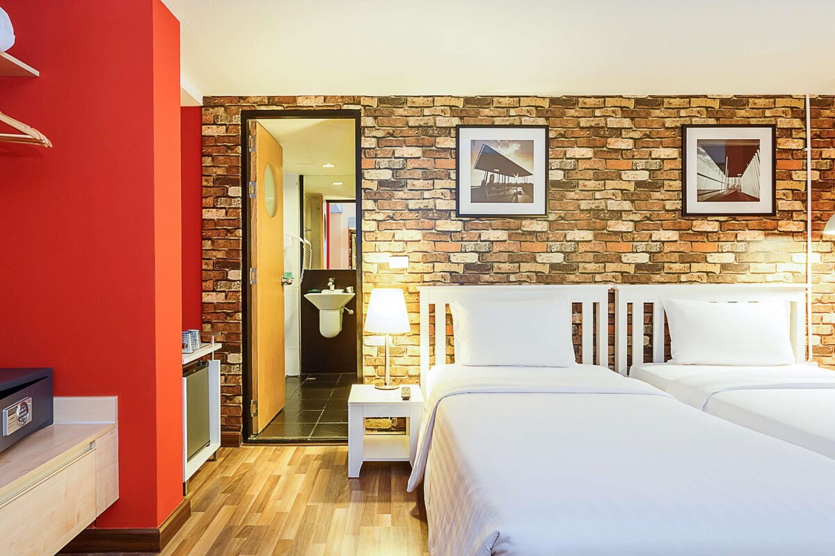 A hotel room with a brick wall, two beds, a small bedside table with a lamp, a bathroom visible through an open door, and a red wall on one side.