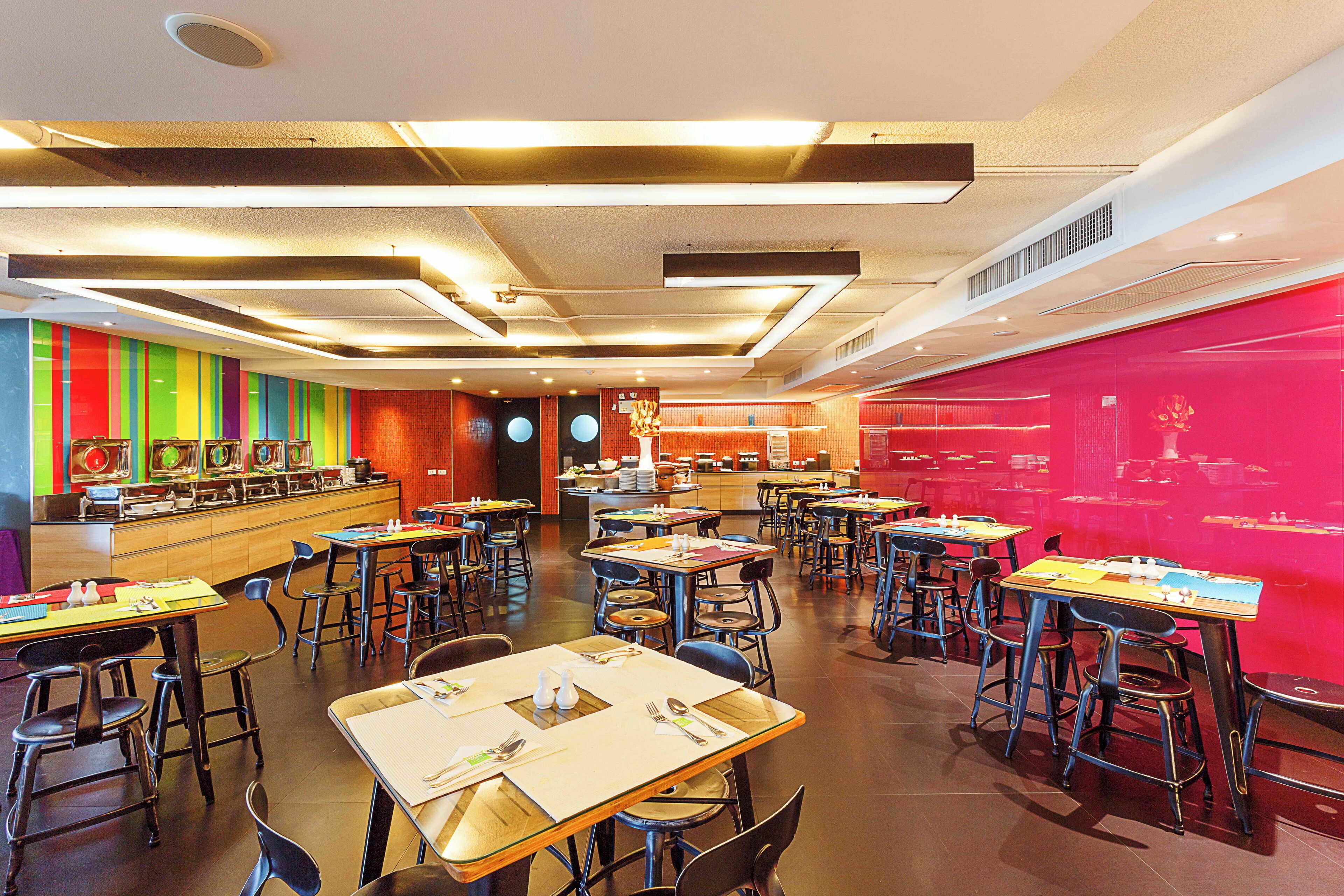 A modern restaurant with multiple dining tables and chairs, a colorful wall, and a counter with food items.