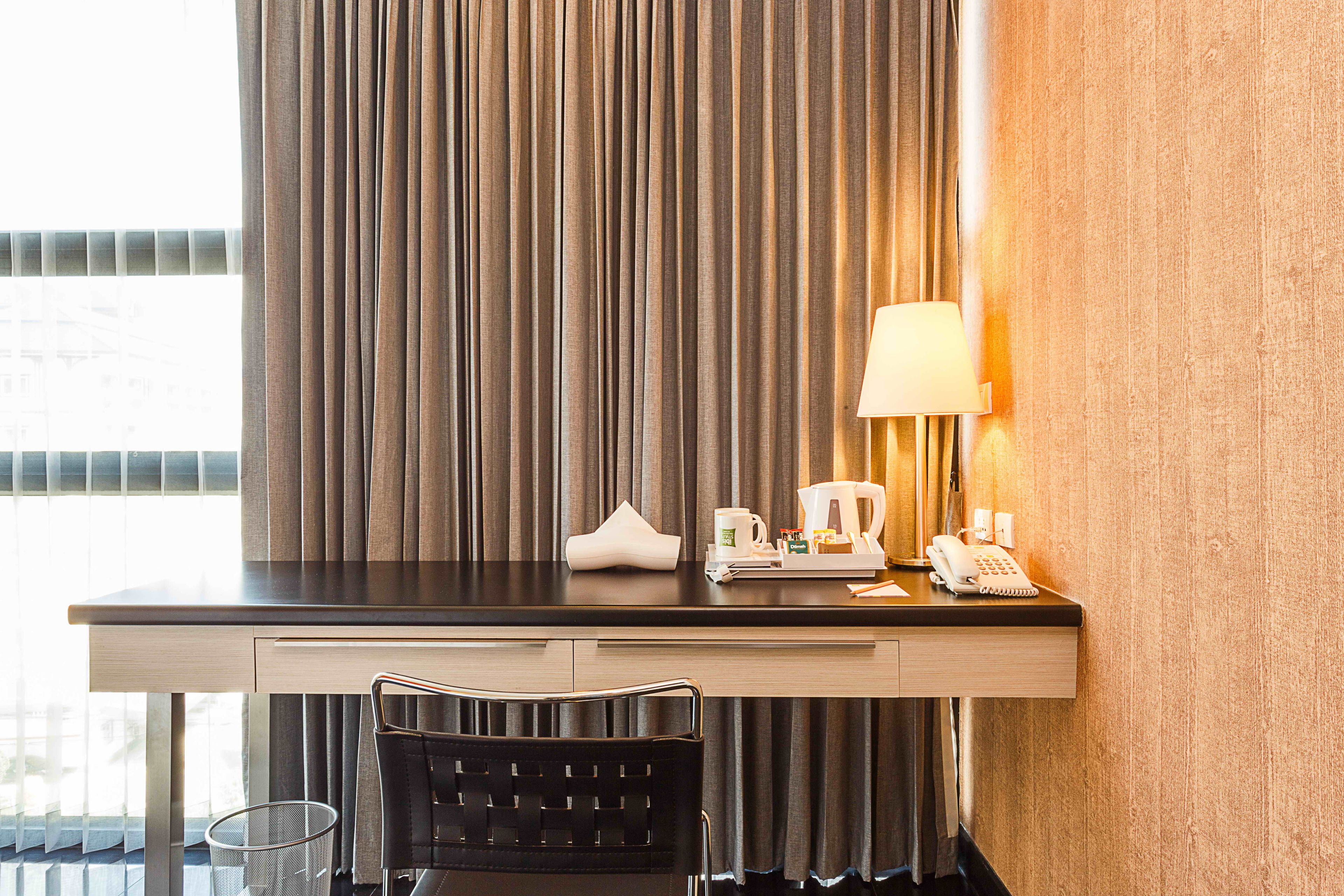Ein Hotelzimmer mit Schreibtisch, Stuhl, Lampe, Telefon, Kaffeemaschine und einem kleinen Tisch mit Tasse und Taschentuch.