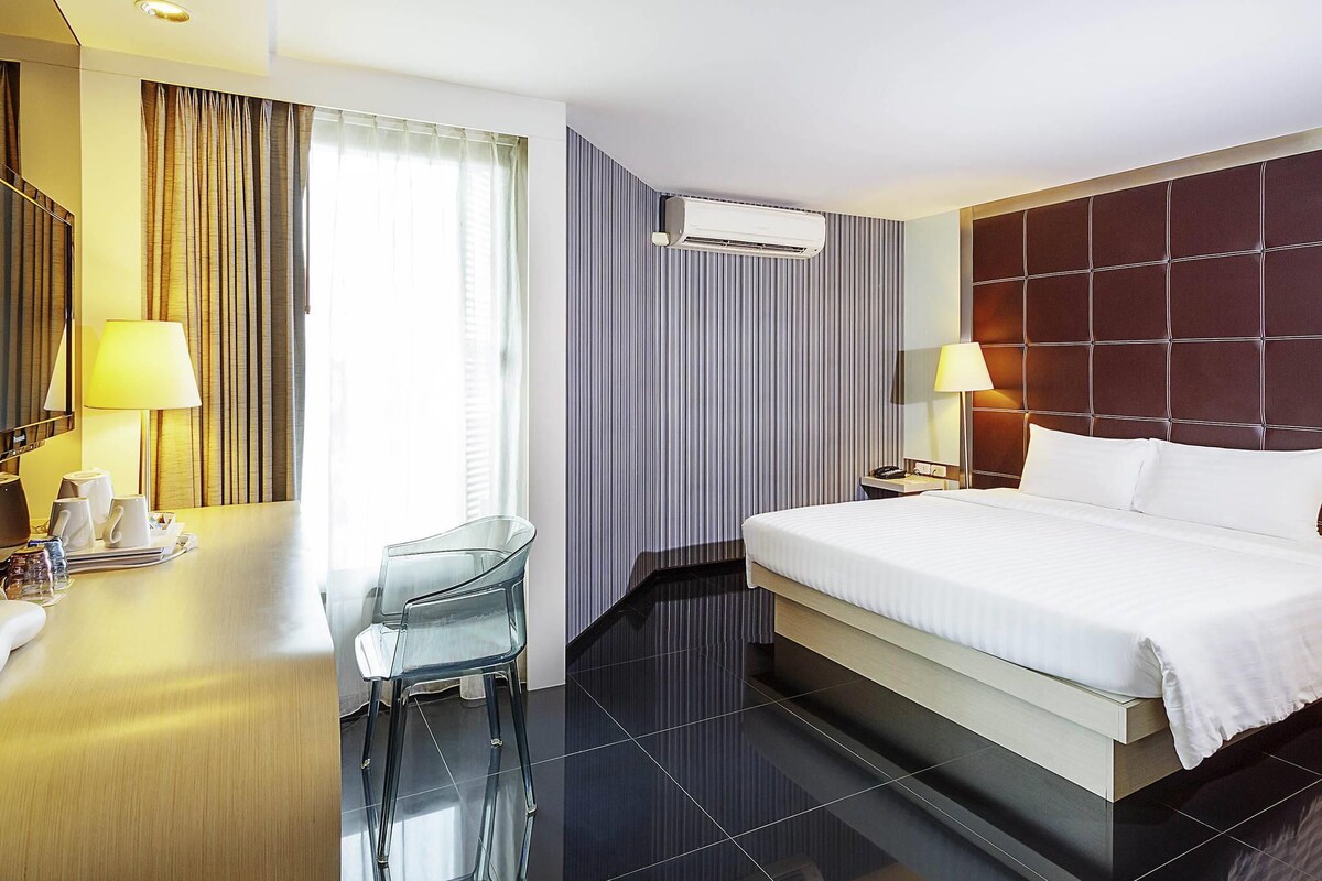 A hotel room with a bed, a desk with a chair, an air conditioning unit, and a window with curtains.