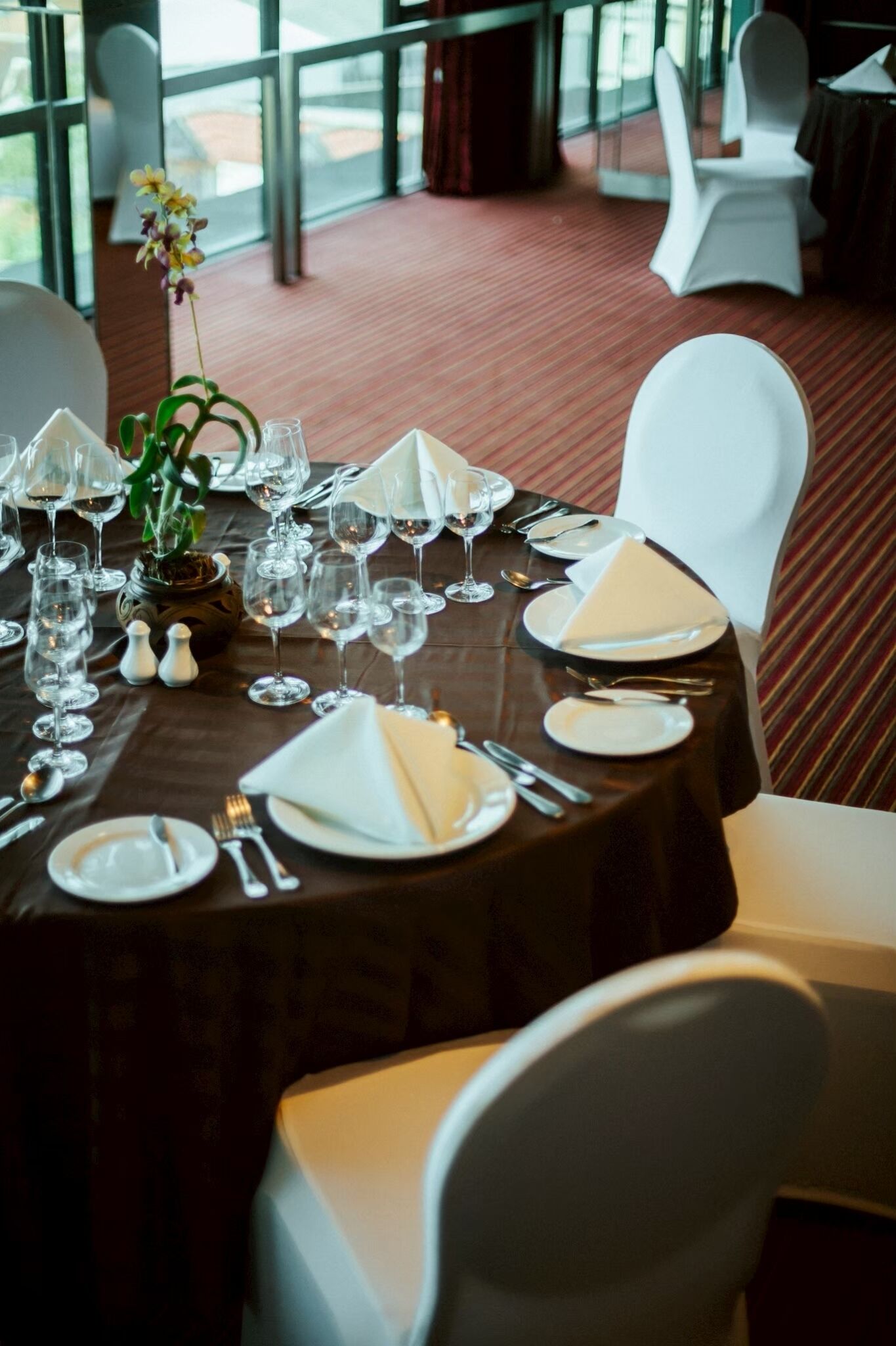 Una mesa de comedor lista para una comida, con sillas blancas, un florero con flores y vistas al exterior a través de amplias ventanas.