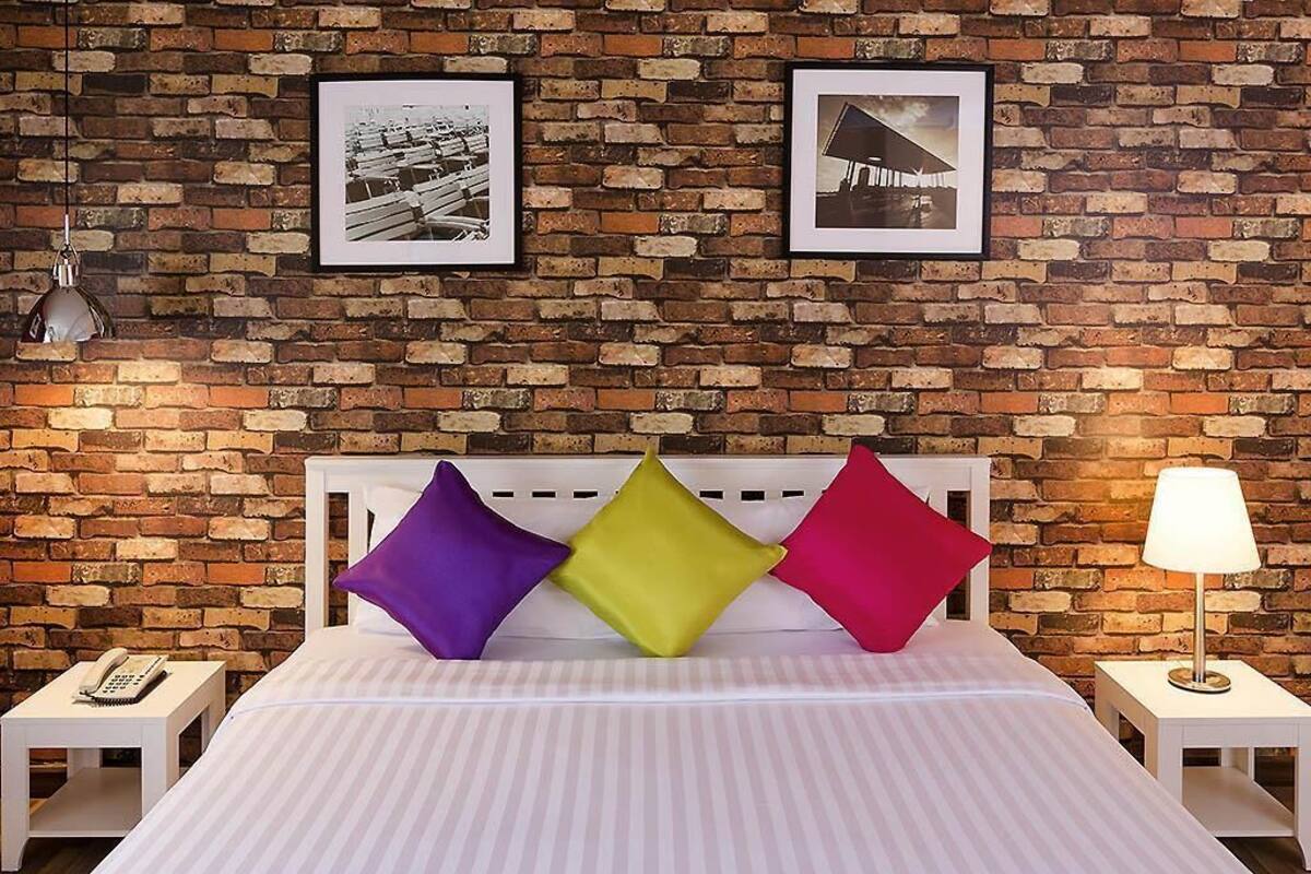 A hotel room with a brick wall, a bed with striped bedding, three pillows, a nightstand with a lamp, and two framed pictures.