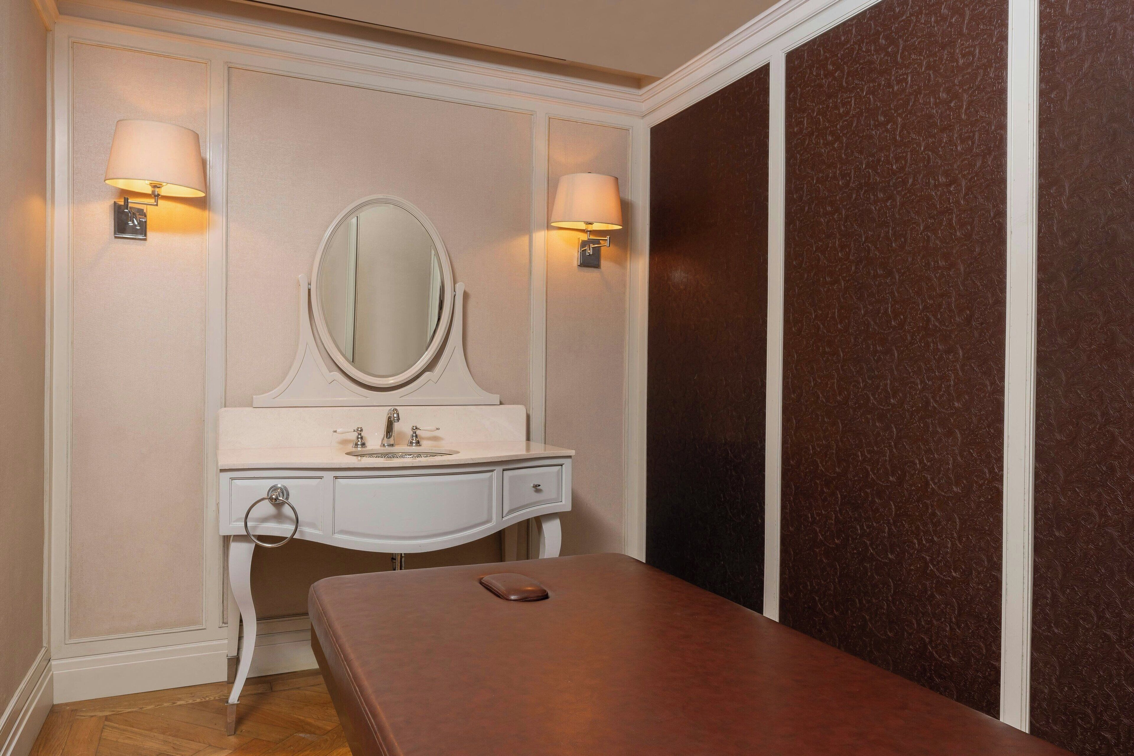 Une pièce comprenant une table de massage en bois, une coiffeuse blanche avec un miroir et deux lampes fixées au mur.