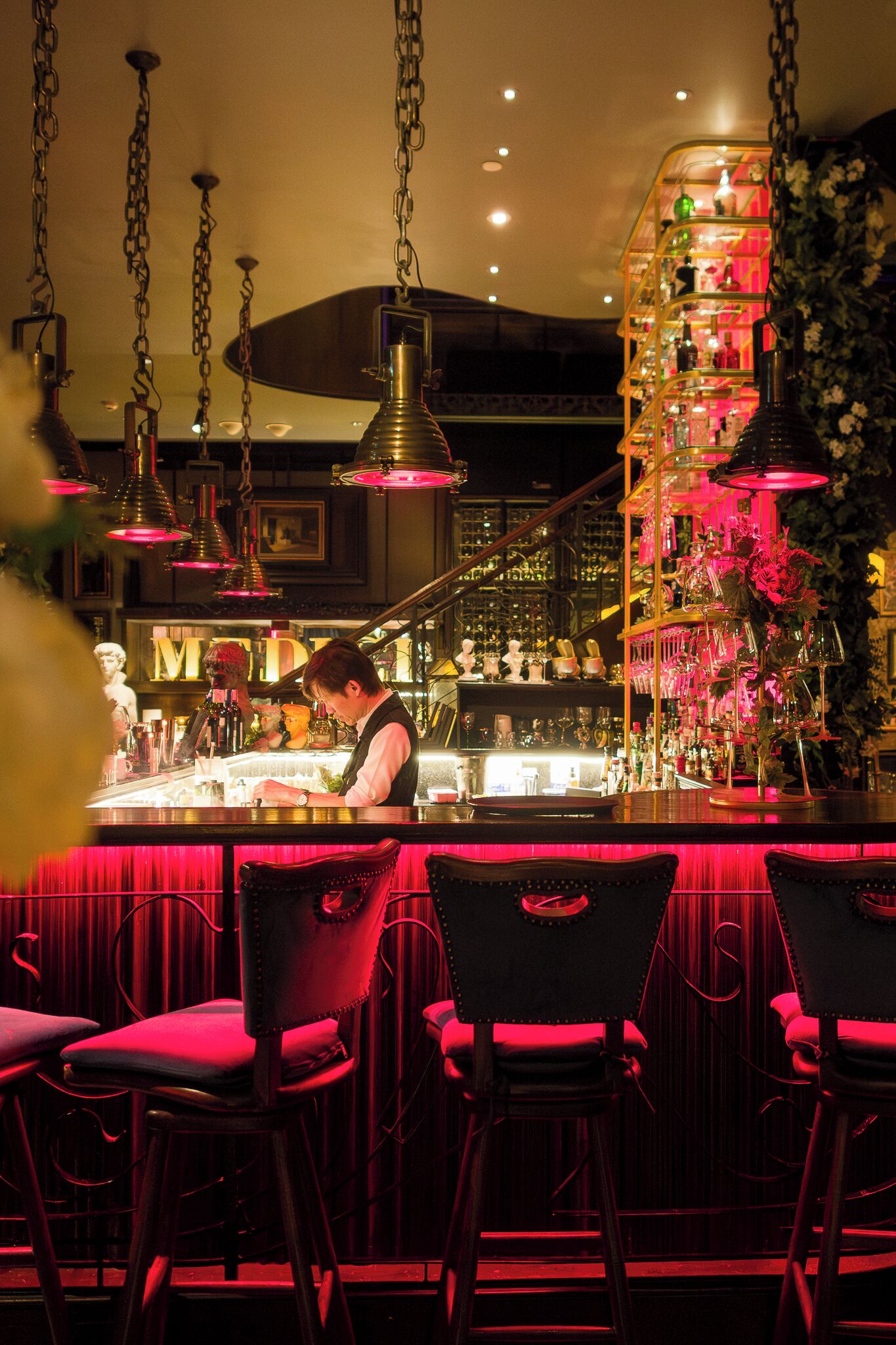 A bar area with a bartender, red bar stools, and a well-lit, decorative shelf with bottles and flowers.