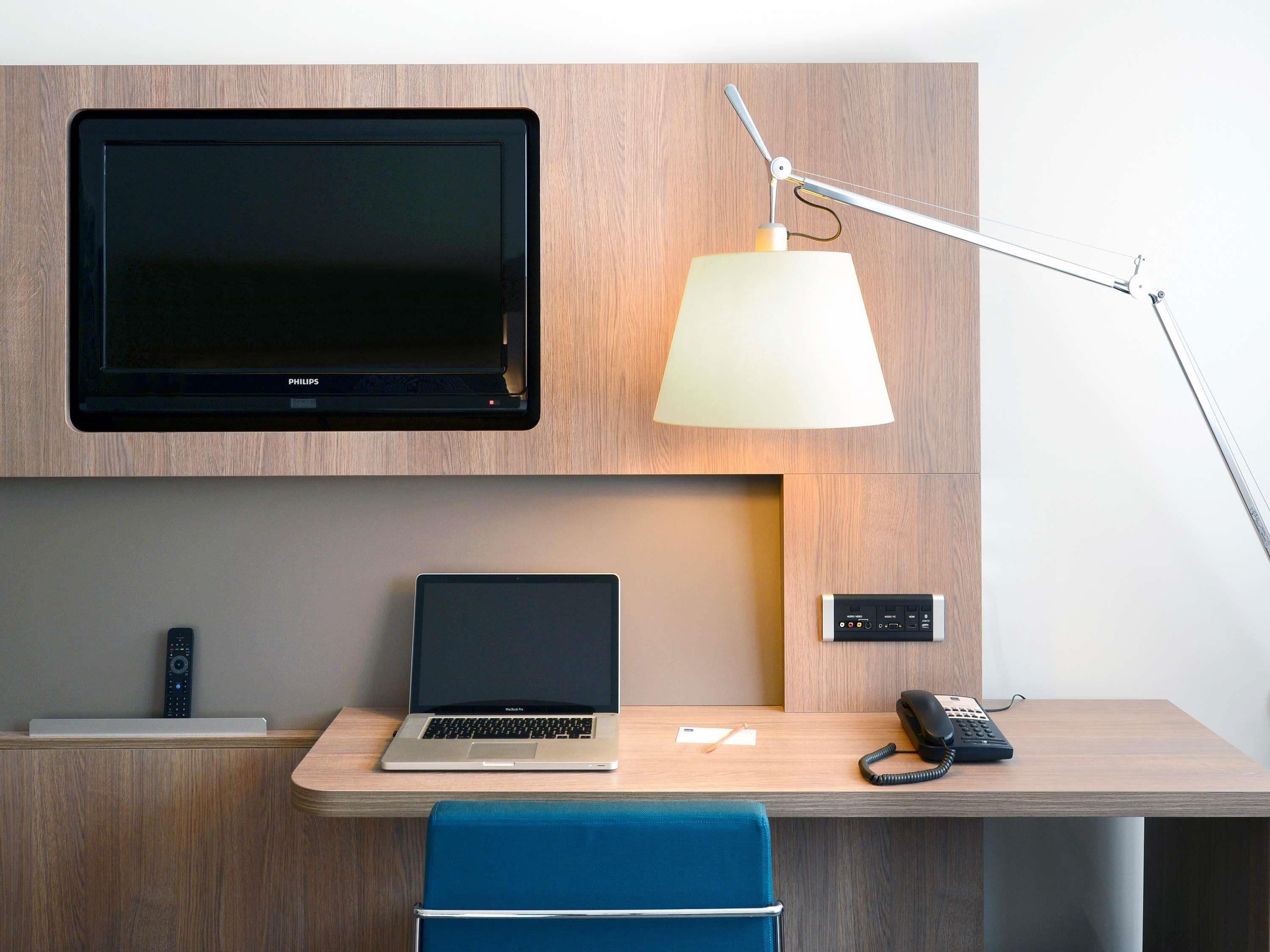 A modern hotel room with a flat-screen TV mounted on the wall, a desk with a laptop, a telephone, and a lamp.