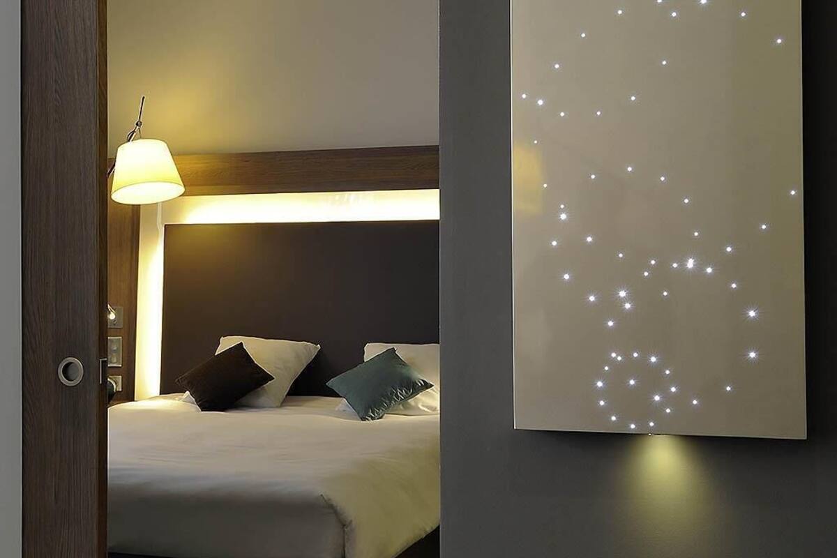 A bedroom with a bed, pillows, a wall-mounted light fixture, and a decorative light panel.